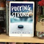 An reader sits on a book shelf showing the cover for novel Pucking Strong that has a hockey puck on an ice rink